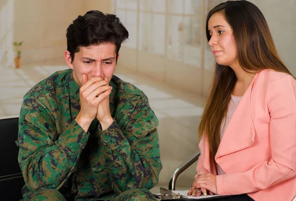 Handsome young soldier wearing uniform suffering from stress, with a woman therapist in a medical health center, covering his mouth using both hands and remembering the horrors of war, in a office — Stock Photo, Image