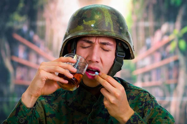 Guapo joven soldado vestido de uniforme que sufre de estrés después de la guerra, sosteniendo en su mano un vaso de ron y comiendo algunas pastillas, en un fondo borroso —  Fotos de Stock