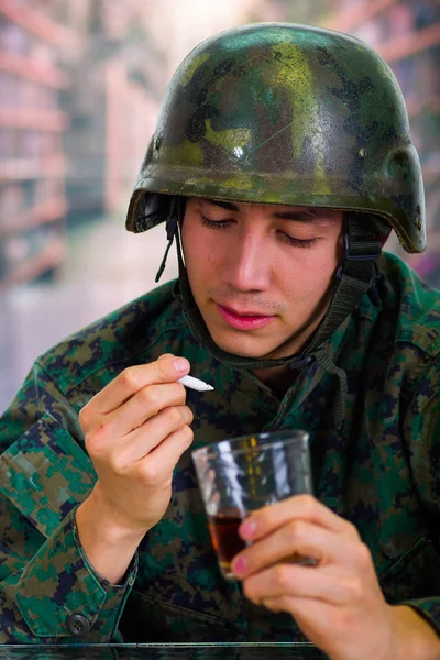 Bonito jovem soldado vestindo uniforme sofrendo de estresse no pós-guerra, segurando em sua mão um copo de ron e um charro de cannabis, em um fundo embaçado — Fotografia de Stock