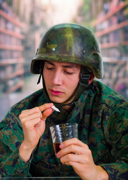 Guapo joven soldado vestido de uniforme que sufre de estrés después de la guerra, sosteniendo en su mano un vaso de ron y un porro de cannabis, en un fondo borroso —  Fotos de Stock