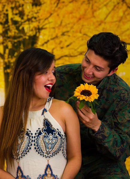 Surprised wife looking at her husband arrived, in army uniform and holding a sunflower as gift, in a yellow park background — Stock Photo, Image