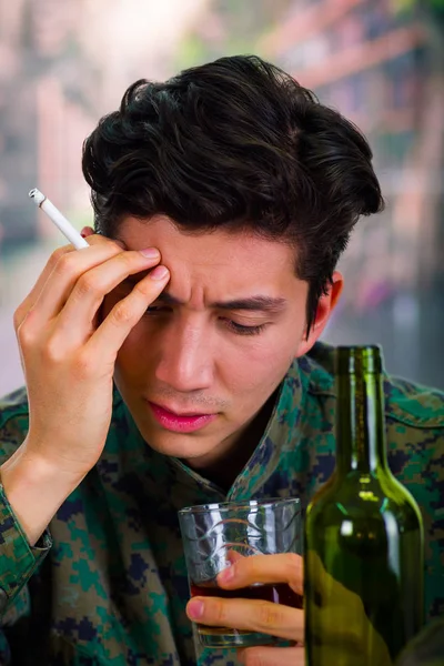 Красиве мислення молодого солдата в уніформі, що страждає від стресу після війни, зі склянкою і пляшкою алкоголю поруч з ним, тримаючи цигарку в руці, на розмитому фоні — стокове фото