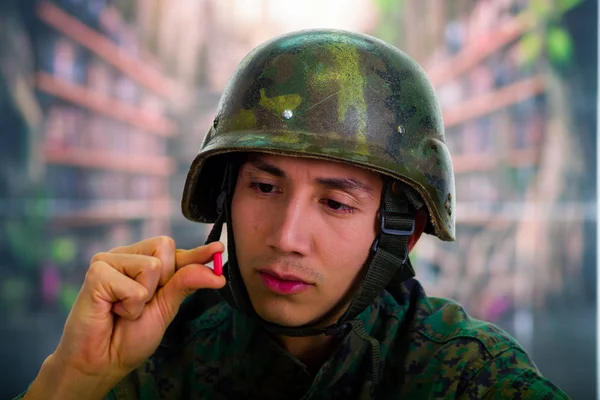 Bonito jovem soldado vestindo uniforme sofrendo de estresse no pós-guerra, segurando em sua mão uma pílula vermelha, em um fundo embaçado — Fotografia de Stock