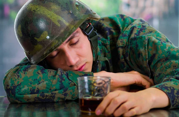 Beau jeune soldat endormi portant l'uniforme souffrant de stress après la guerre, tenant dans sa main un verre de ron, dans un fond flou — Photo