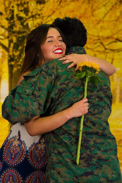 Portrait of happy wife holding a sunflower and hugging her husband in army uniform, in a yellow park background — Stock Photo, Image