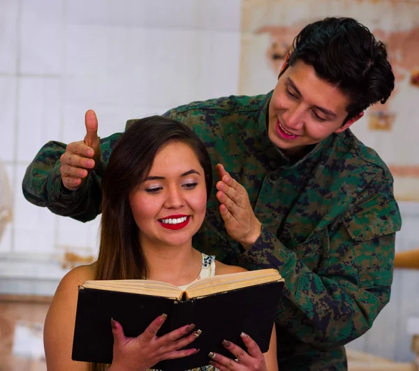 Close up de um homem vestindo um uniforme do exército, surpreendendo sua esposa enquanto ela está lendo um livro, em um fundo branco borrado — Fotografia de Stock