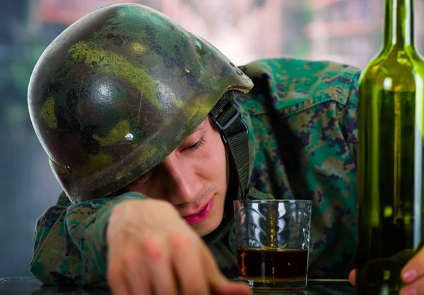 Guapo joven soldado vestido de uniforme que sufre de estrés después de la guerra, con un vaso de ron y botella a su lado, en un fondo borroso — Foto de Stock