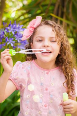 Bir yaz niteliğine sabun köpüğü ile oynarken, bir bulanık doğa arka planda kafasının içinde bir pembe elbise ve çiçek giyen mutlu küçük kıvırcık kız