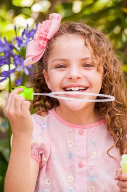 Bir yaz niteliğine sabun köpüğü ile oynarken, bir bulanık doğa arka planda kafasının içinde bir pembe elbise ve çiçek giyen mutlu küçük kıvırcık kız