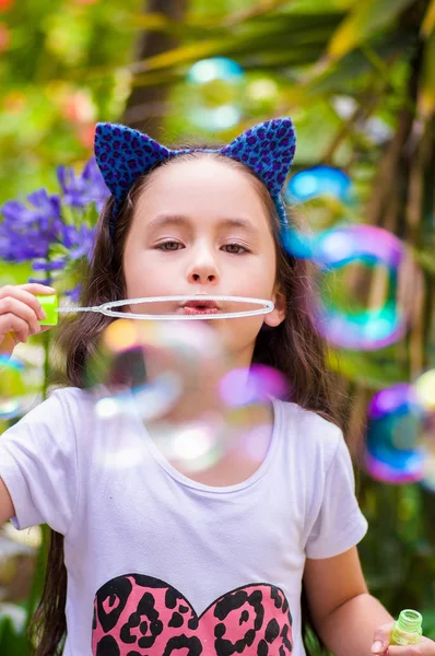 Glückliches kleines Mädchen spielt mit Seifenblasen auf einem Sommer-Natur, trägt ein blaues Ohren Tiger Accessoires über dem Kopf in einem verschwommenen Natur Hintergrund — Stockfoto