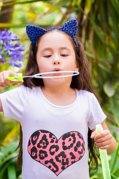 Glückliches kleines Mädchen spielt mit Seifenblasen auf einem Sommer-Natur, trägt ein blaues Ohren Tiger Accessoires über dem Kopf in einem verschwommenen Natur Hintergrund — Stockfoto