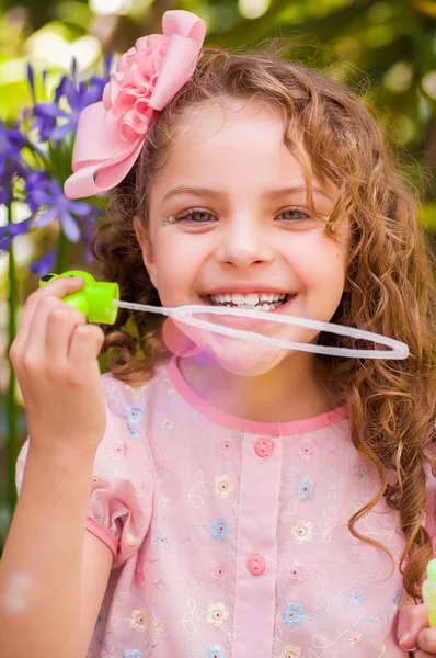 Bir yaz niteliğine sabun köpüğü ile oynarken, bir bulanık doğa arka planda kafasının içinde bir pembe elbise ve çiçek giyen mutlu küçük kıvırcık kız — Stok fotoğraf