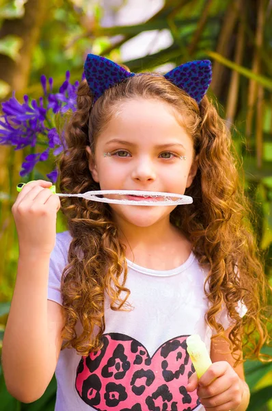 Glad liten lockigt flicka leker med såpbubblor på en sommar natur, klädd i en blå öron av tiger tillbehör över hennes huvud i en suddig natur bakgrund — Stockfoto