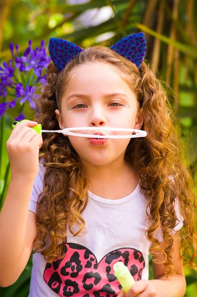 Blij krullend meisje spelen met zeepbellen op een zomer natuur, het dragen van een blauwe oren van tijger accessoires over haar hoofd op een achtergrond wazig natuur — Stockfoto