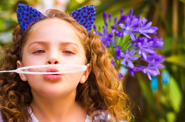 Portrait d'une heureuse petite fille bouclée jouant avec des bulles de savon sur une nature estivale, portant une oreille bleue d'accessoires de tigre sur sa tête dans un fond de nature floue — Photo