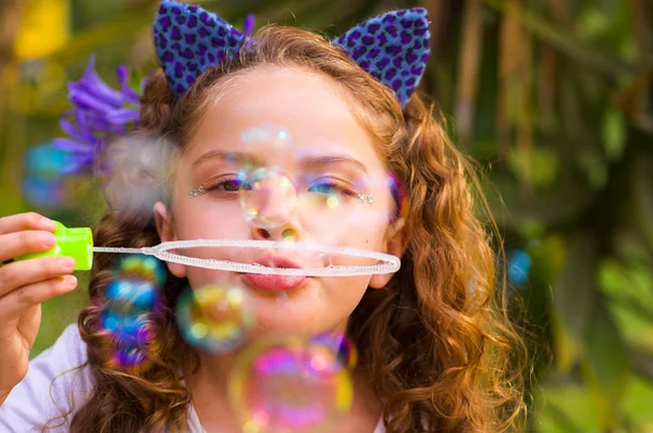 Porträt eines glücklichen kleinen lockigen Mädchens, das mit Seifenblasen in der sommerlichen Natur spielt und blaue Ohren mit Tiger-Accessoires über dem Kopf trägt, vor verschwommenem Naturhintergrund — Stockfoto