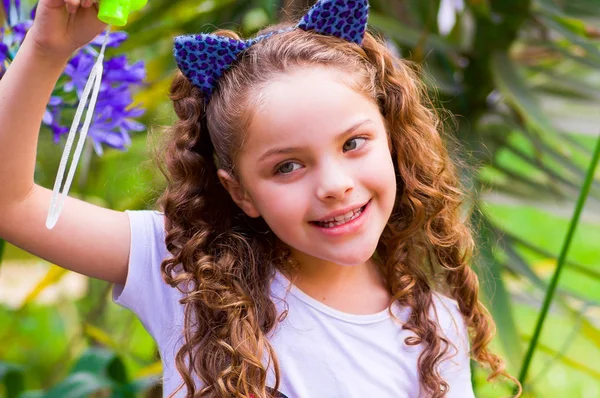 Blij krullend lachende meisje spelen met zeepbellen op een zomer natuur, het dragen van een blauwe oren van tijger accessoires over haar hoofd op een achtergrond wazig natuur — Stockfoto