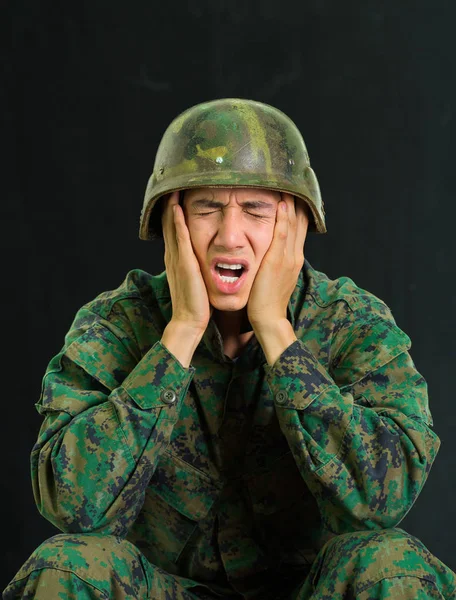 Красивий молодий солдат у формі, що страждає від стресу, кричить закритими очима і використовує обидві руки, щоб торкнутися його щоки, на чорному тлі — стокове фото