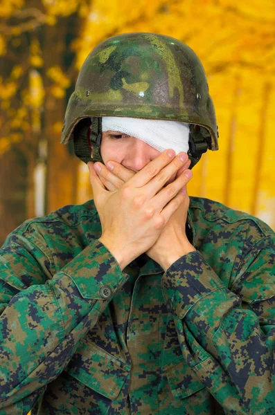 Красивий молодий солдат у формі, що страждає стресом, з білою пов'язкою навколо голови і прикриває око, обидва прикриваючи рот, на розмитому жовтому фоні — стокове фото