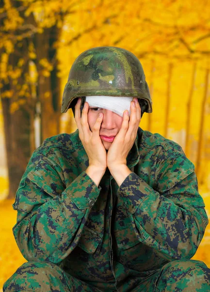 Guapo joven soldado vestido de uniforme que sufre de estrés, con un vendaje blanco alrededor de la cabeza y cubriendo su ojo, con ambas manos en las mejillas, en un fondo amarillo borroso — Foto de Stock
