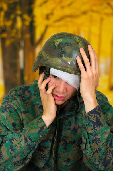 Bonito jovem soldado vestindo uniforme sofrendo de estresse, com uma ligadura branca em torno de sua cabeça e cobrindo o olho, tocando com uma mão o capacete e cobrindo seu rosto com a outra mão, em — Fotografia de Stock