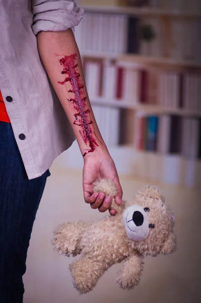 Close up of a young depressive woman with her arm bleeding, holding in her hand a teddy bear, in a blurred background — Stock Photo, Image