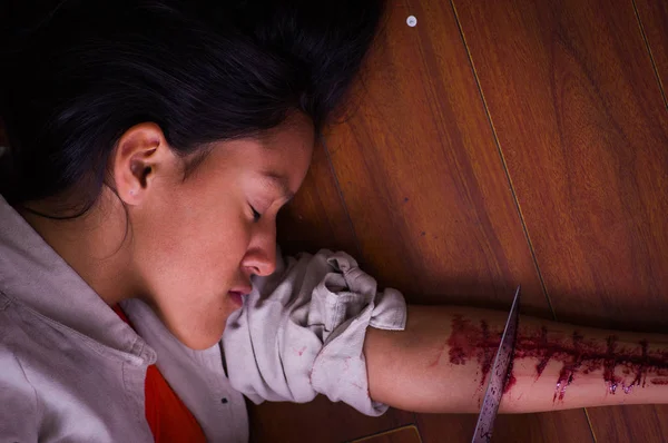 Primo piano di una giovane donna depressiva che tiene in mano un coltello, con il braccio sanguinante, disteso a terra, su uno sfondo di legno — Foto Stock