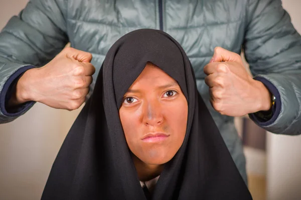 Portrait of a beautiful muslim girl wearing a hijab, with a white man behind her using both hands pretending to hit her head, in a blurred background