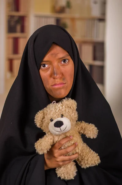 Portret van een trieste moslim meisje het dragen van een hijab en houden haar armen haar teddybeer, in een onscherpe achtergrond — Stockfoto
