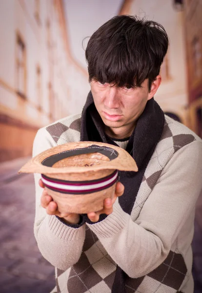 Close up of a sad homeless young man in the streets, with a hat in his hands, asking for money, in a blurred background — Stock Photo, Image