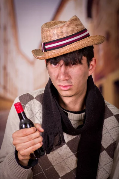 Close up of a sad homeless young man in the streets, wearing a hat and a scarf, and holding a bottle of wine in his hand, in a blurred background — Stock Photo, Image