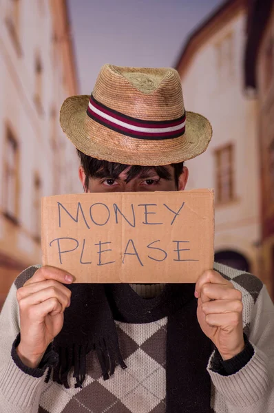 Close up of a homeless with cardboard description of money please, wearing a hat, in a blurred background
