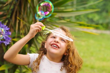 Bir yaz niteliğine sabun köpüğü ile oynarken, bir bulanık doğa arka planda kafasının içinde bir pembe elbise ve çiçek giyen mutlu küçük kıvırcık kız