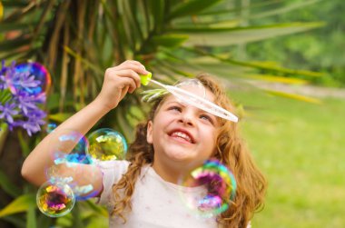 Bir yaz niteliğine sabun köpüğü ile oynarken, bir bulanık doğa arka planda kafasının içinde bir pembe elbise ve çiçek giyen mutlu küçük kıvırcık kız