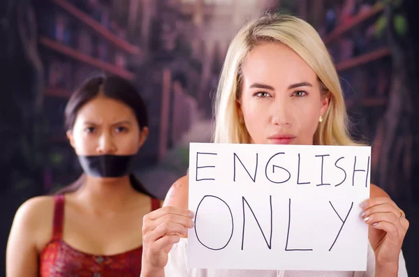 Young mad american blonde woman holding a piece of paper with a description of english only, while latin is with a black tape in her mouth, racism, violence or discrimination concept