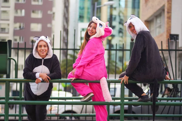 Close up of a happy group of friends, waiting the public transportation and wearing different costumes, one woman wearing a pink unicorn costume, other woman a panda costume and the man wearing a cat
