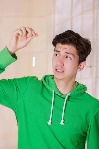 Hombre guapo con una cara sorprendida sosteniendo un tampón de algodón de menstruación, con una sudadera con capucha verde en un fondo borroso — Foto de Stock