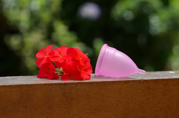 Feminine hygiene product - Menstrual cup over a wooden structure next to a beautiful red flowers, in a blurred background — Stock Photo, Image