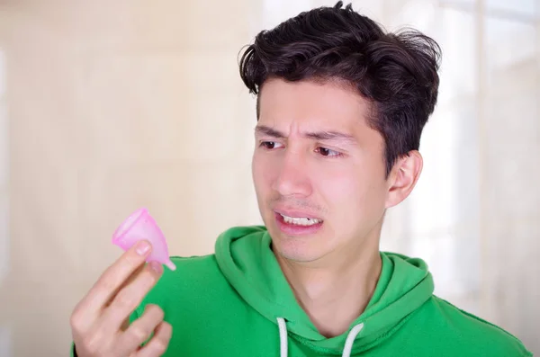 Close up de um homem fazendo um rosto nojento enquanto está segurando um copo menstrual em suas mãos, em um fundo borrado — Fotografia de Stock