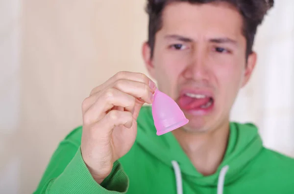 Close up of a man doing an disgusting face while is holding a menstrual cup in his hands, in a blurred background — Stock Photo, Image