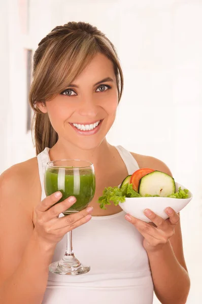 Vackra unga leende kvinna bär en vit t-shirt och håller en frisk sallad med ena handen och glas juice i hennes andra hand — Stockfoto