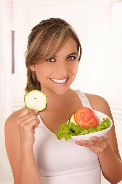 Hermosa joven vistiendo una camiseta blanca y sosteniendo una ensalada saludable con una mano y un trozo de pepino en su otra mano — Foto de Stock