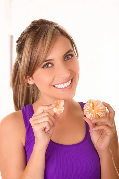 Bela jovem mulher sorridente vestindo uma camisa roxa e comendo uma tangerina em um fundo branco — Fotografia de Stock