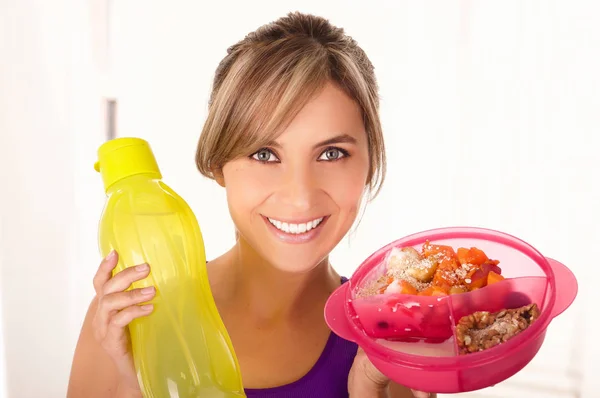 Mooie lachende vrouw die een paarse t-shirt en houden van een gezonde fruitsalade in één hand en een gele fles water in een witte achtergrond — Stockfoto