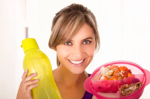 Mulher sorridente bonita vestindo uma camiseta roxa e segurando uma salada de frutas saudável em uma mão e uma garrafa de água amarela em um fundo branco — Fotografia de Stock