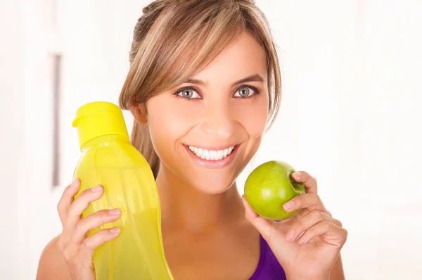 Mulher sorridente bonita vestindo uma camiseta roxa e segurando uma garrafa amarela de água e uma maçã verde em sua outra mão em fundo branco — Fotografia de Stock