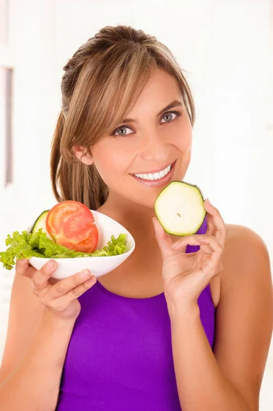 Mooie vrouw die een paarse t-shirt en bezit is van een gezonde salade en een stukje komkommer in haar andere hand op witte achtergrond — Stockfoto