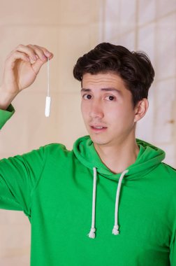 Bulanık bir arka planda yeşil kapüşonlu kameraya bakarak ve bir adet pamuk tampon tutan yakışıklı adam