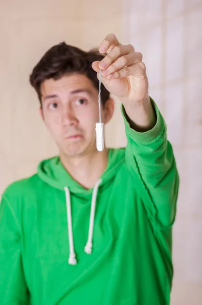 Handsome man with a funny face holding a menstruation cotton tampon in front of him, wearing a green hoodie in a blurred background — Stock Photo, Image