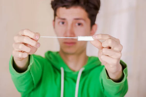 Bonito homem com um rosto engraçado segurando um tampão de algodão menstruação na frente dele, usando um capuz verde em um fundo borrado — Fotografia de Stock